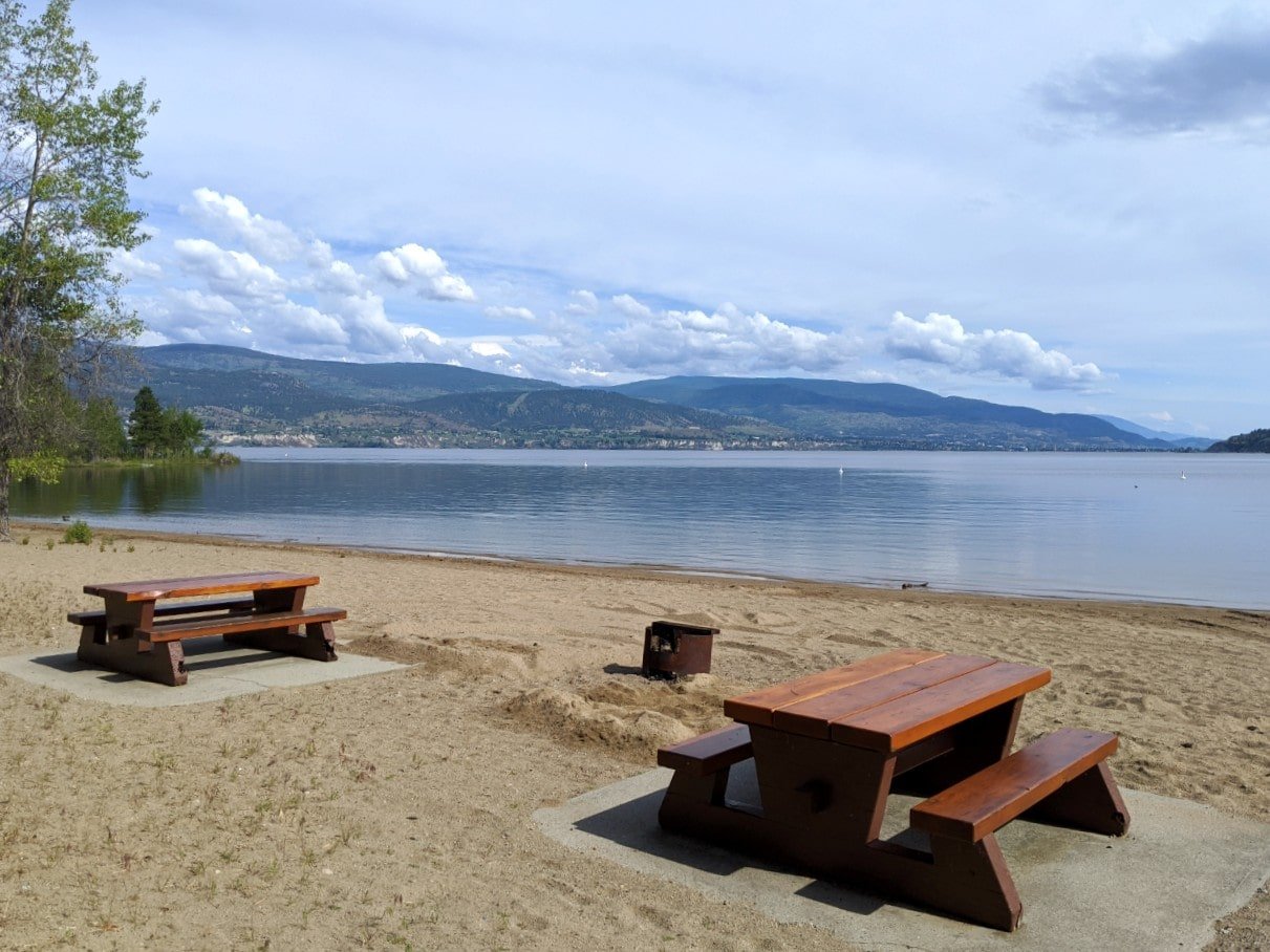 Two picnic tables on sandy beach with firepit next to calm lake and Naramata and Penticton beyond