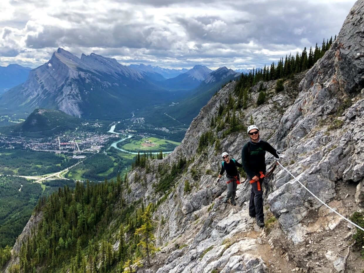 JR and Gemma on Via Ferrata course attached to rock with views of Banff and Mount Rundle behind