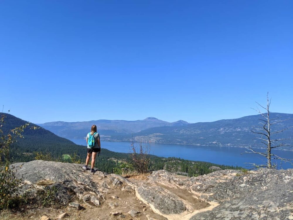 JR stands with her back to camera on a rock looking out over the views of Okanagan Lake and the surrounding hills at Predator Ridge
