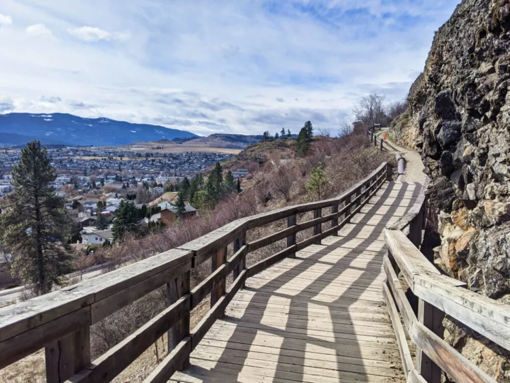 Boardwalk path around side of rocky mountain, with views of Vernon in background