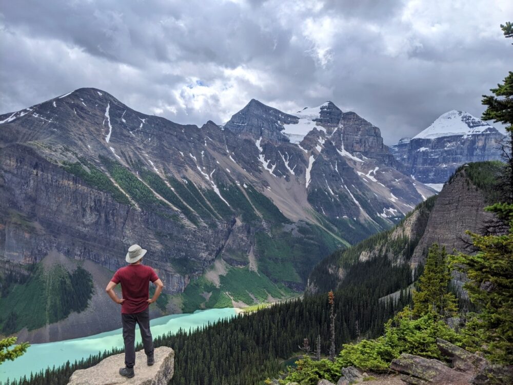 JR standing with back to camera looking down on bright Lake Louise, lined by snow capped mountains