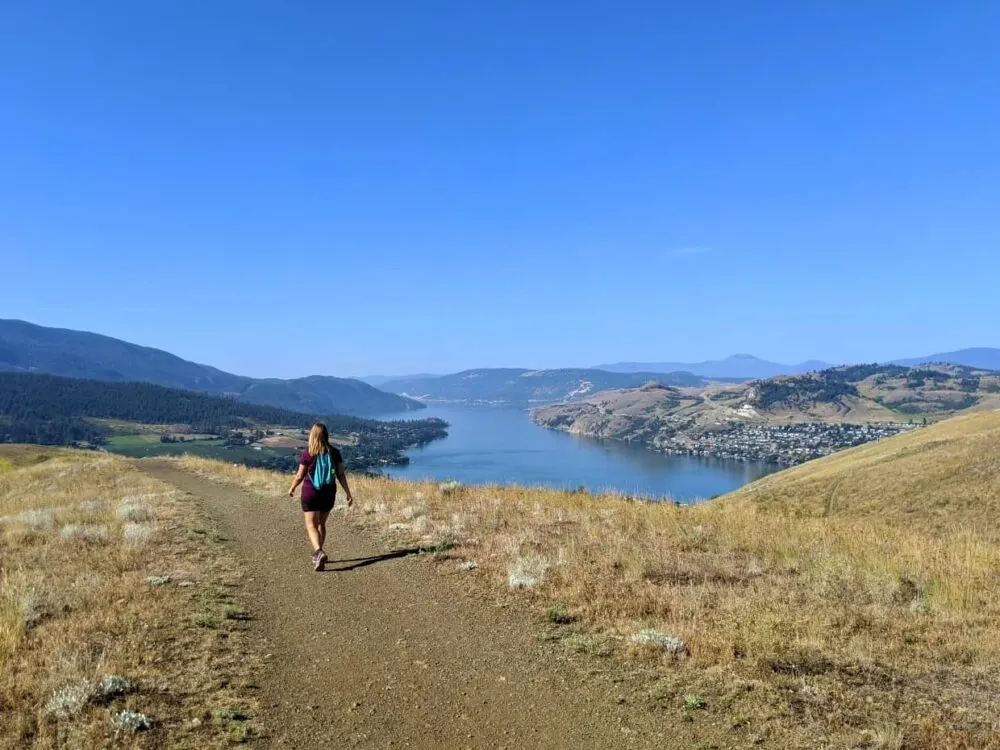 Back view of Gemma walking away along hiking trail with Kalamalka Lake in background, surrounded by rolling hills