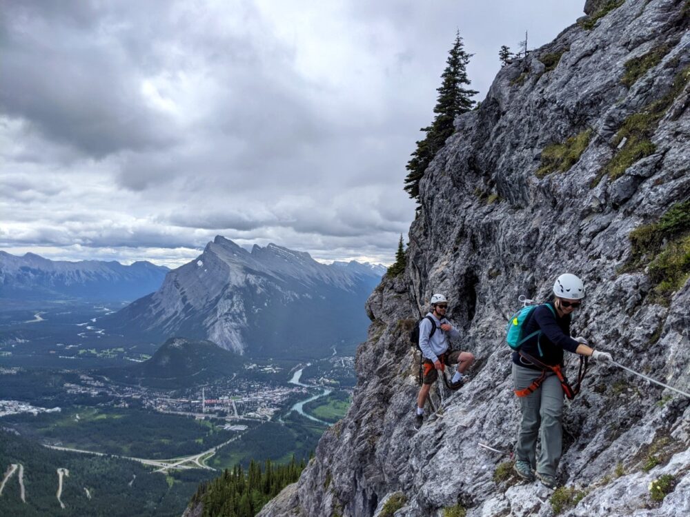 Two people on Via Ferrata course in Banff, holding onto wire on rock to the right, with views of Banff and surrounding mountains in background