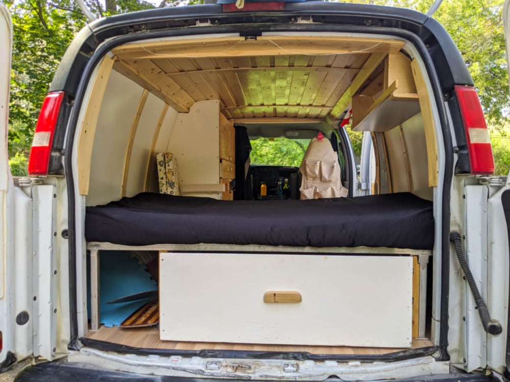 Back view of van with wooden ceiling, mattress on bed, white panel walls, large underbed drawer