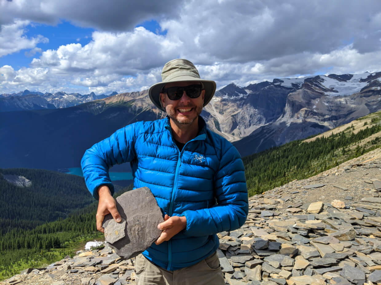 JR smiling and holding up a couple of Burgess Shale fossils to camera at Walcott Quarry, with mountainous background