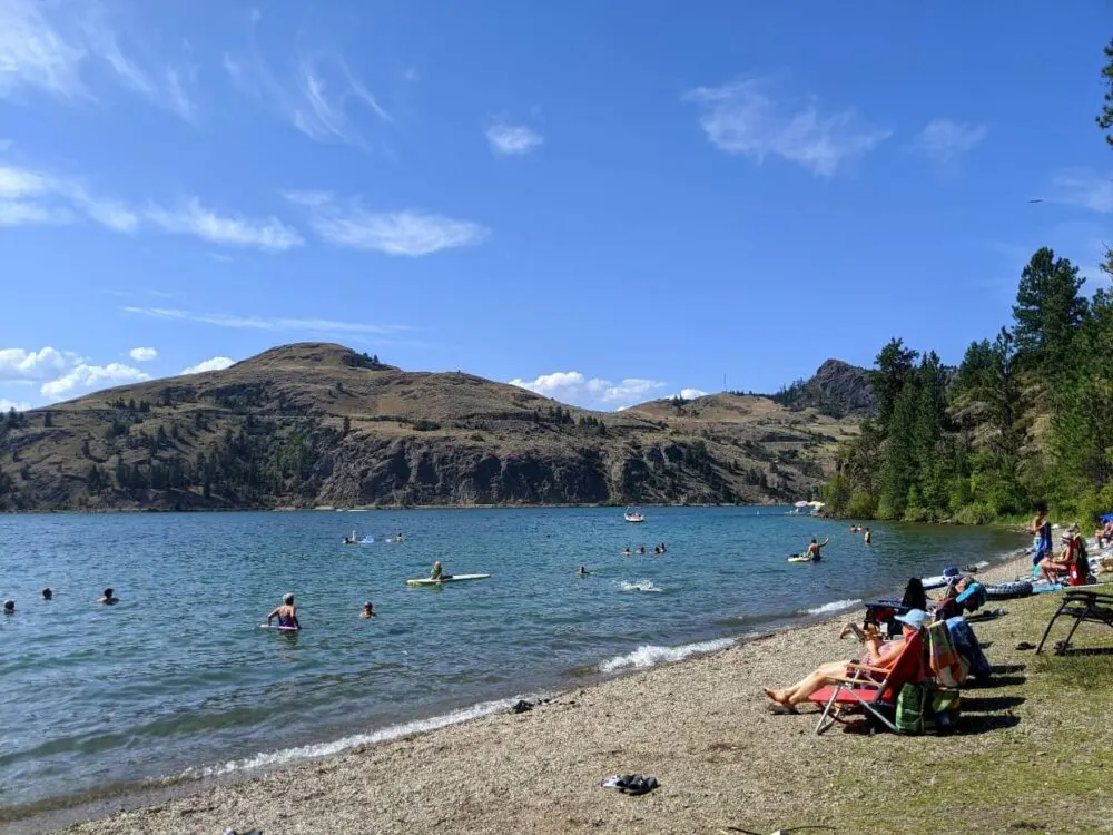 Lakeshore beach in Kalamalka Lake Provincial Park in Vernon, with people sunbathing on beach and swimmers in lake