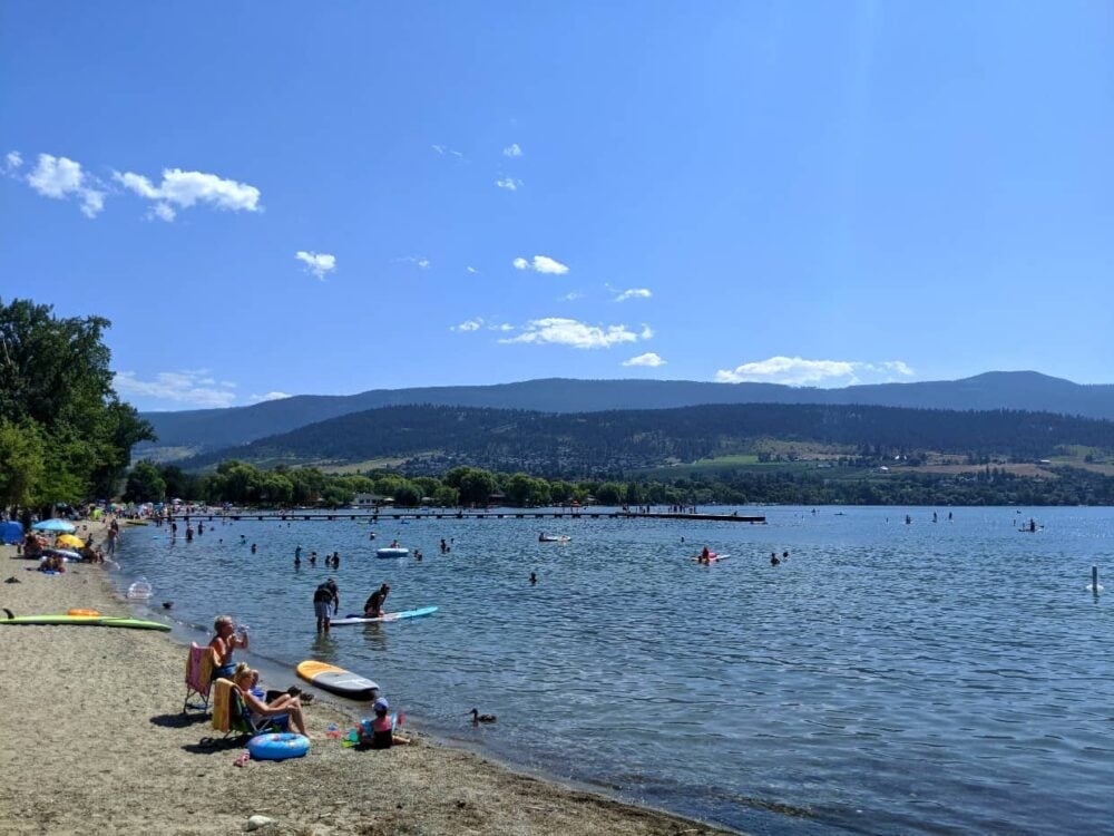 People hang out in chairs and with paddleboards on sandy Kal Beach next to calm Kalamalka Lake in Vernon