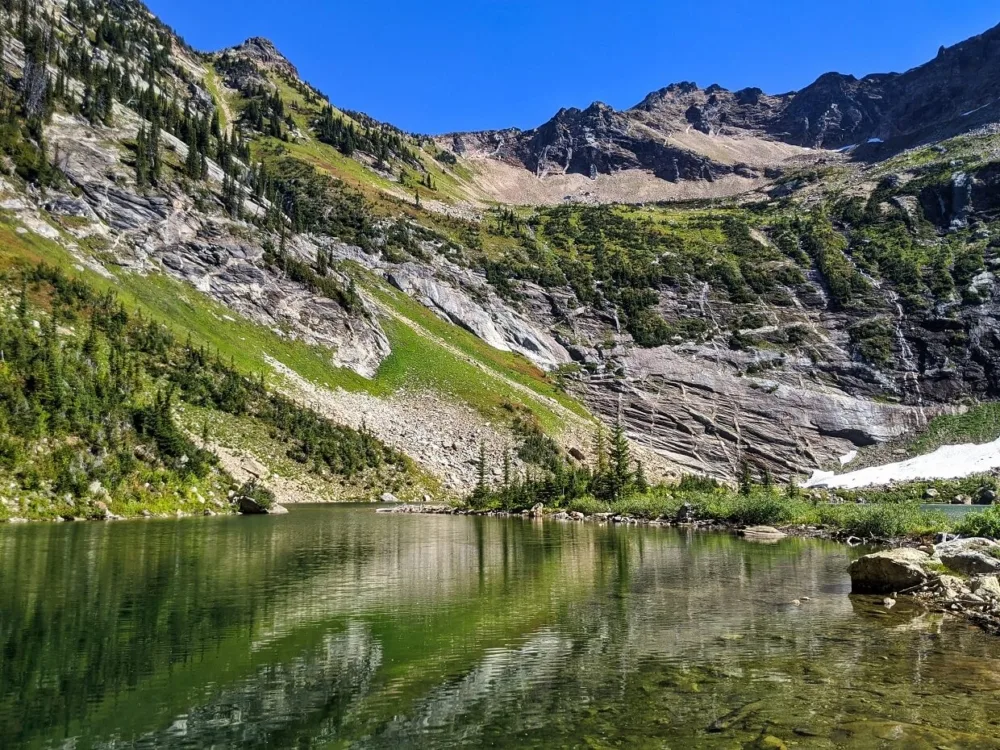 Green coloured reflective lake surrounded by rock cirque, with small waterfalls cascading down slopes, with trees dotted on other slopes