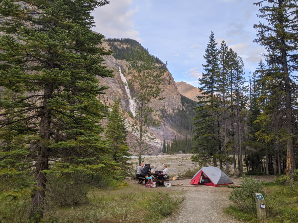A set up tent in a walk-in campsite, with a huge waterfall in the background. The site is surrounded by scattered trees