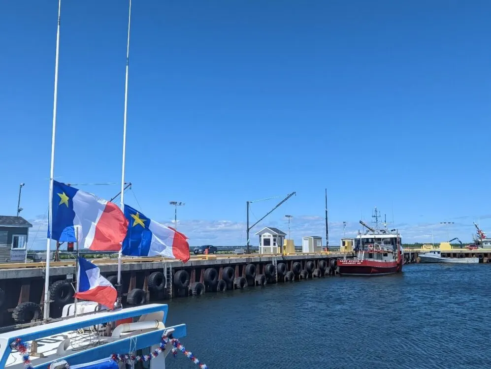 Shippagan marina view with boat sporting Acadian flags on left. There is a red boat visible in the background, docked next to the walkway