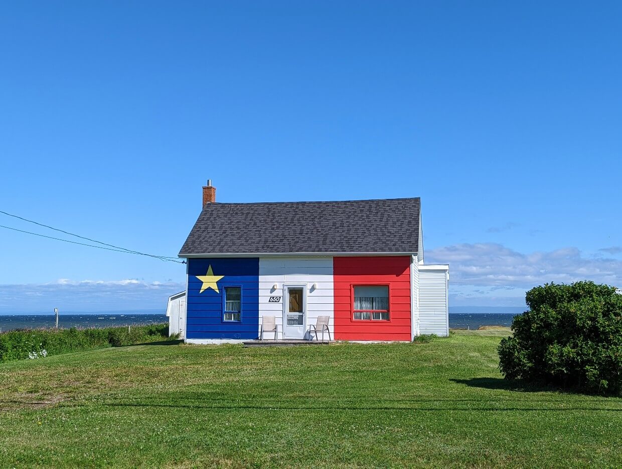 Small brightly coloured house in front of the ocean, painted in the colours of the Acadian flag