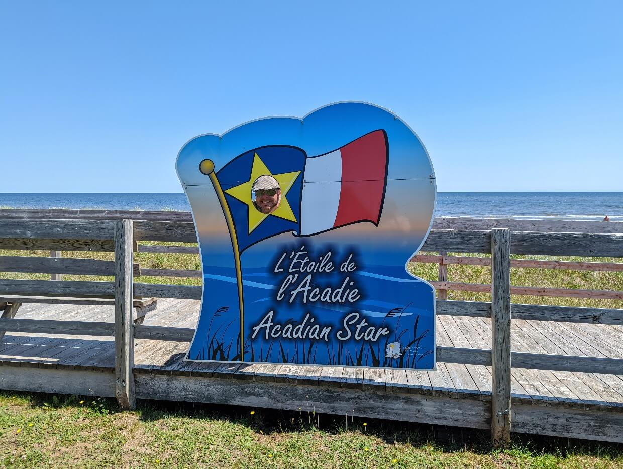Close up of L'Étoilede l'Acadie (Acadian Star) sign with cut out portion on yellow star. JR has put his head through the cut out. The ocean is visible in the background