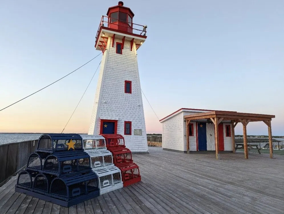 White and red lighthouse on decking with lobster pots painted in Acadian flag colours (blue, white, red and yellow star on blue)