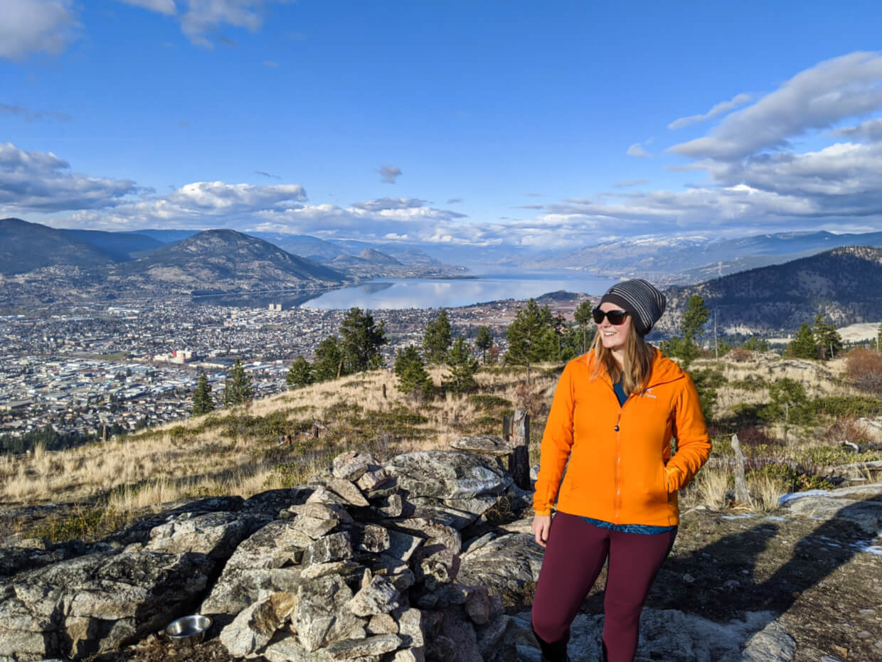 Gemma standing by pile of rocks at top of mountain summit with view of Penticton city centre behind with lake and surrounding mountains