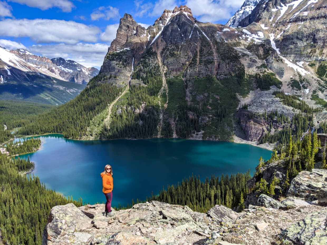 Gemma standing on rock in front of stunning view of blue Lake O'Hara below, surrounded by soaring mountains and dense forest