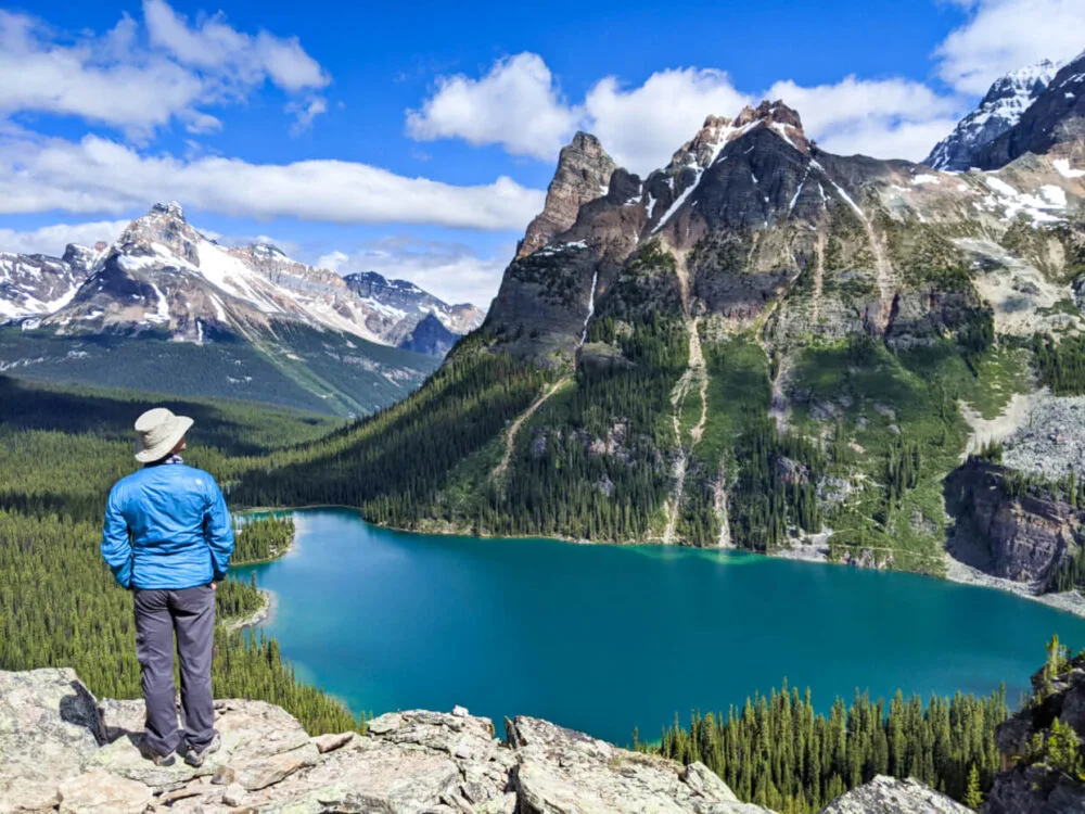 Back view of JR standing on rock looking out to elevated view of rugged mountains and turquoise lake below (Lake O'Hara)