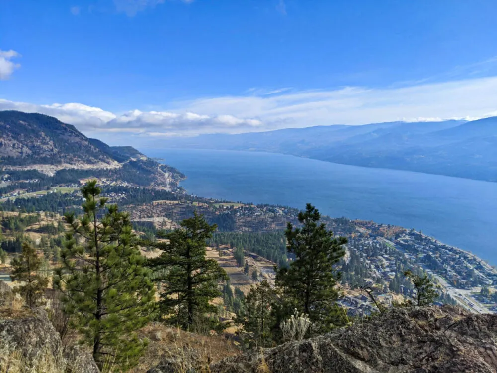 Elevated view looking over trees down to town of Peachland below, next to the shore of Okanagan Lake. The mountains on the other side of the lake are visible