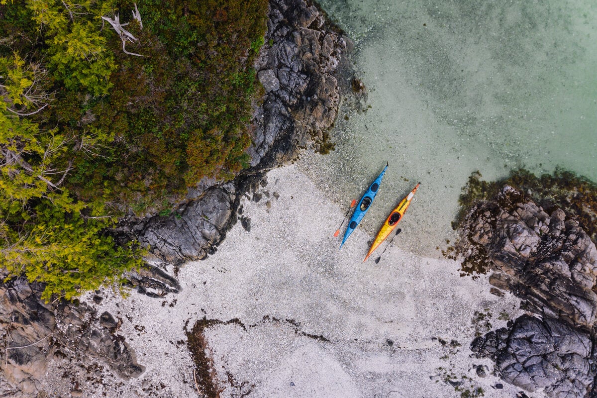 Top down view of kayaks on shore, landing on one of the islands in the Broken Group. Light coloured rock beach with clear ocean water, overhanging trees