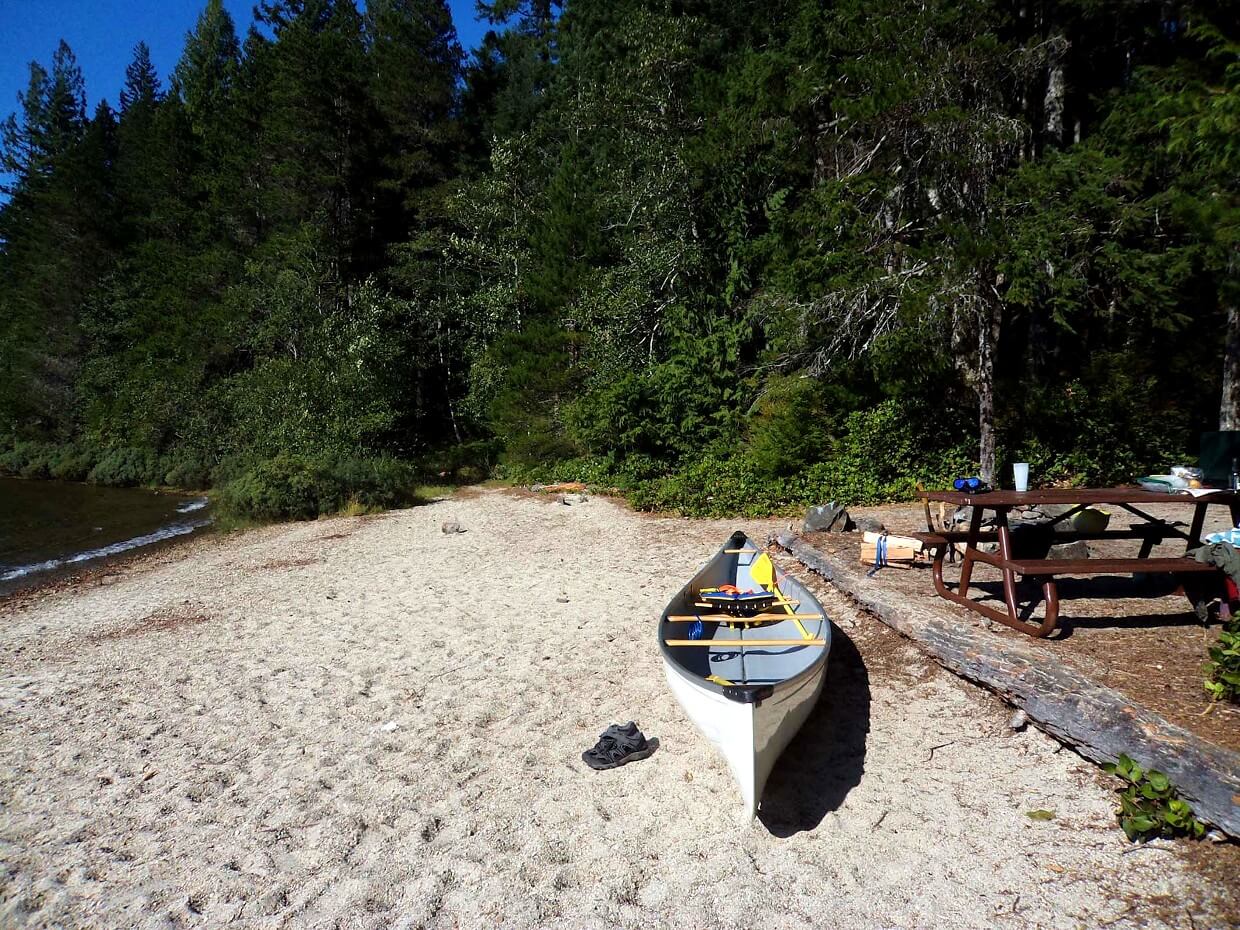 White canoe sits on sandy beach next to calm lake, with picnic bench on right hand side, surrounded by forest