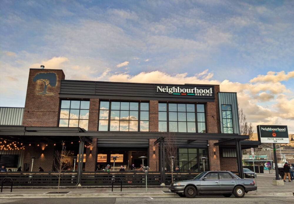 Side view of two floor modern brick building with Neighbourhood Brewing logo on the side. A car is parked on the side of the street on front of the building