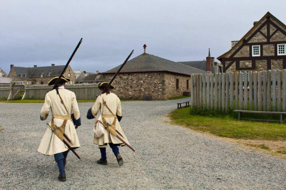 Two Fortress of Louisbourg soldiers (in 18th century clothing) walk away from camera with fortress buildings behind