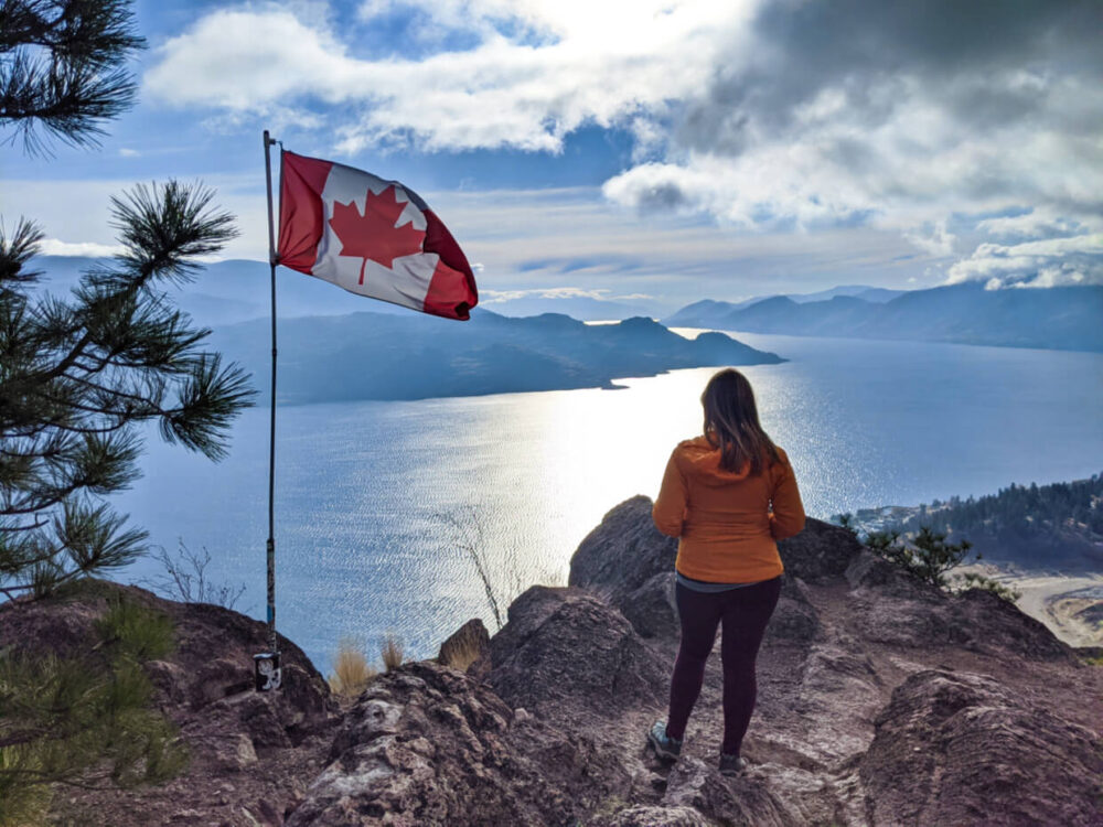 Back view of Gemma taking in the views at the top of Pincushion Mountain. There is a Canadian flag to the left. Okanagan Lake is visible below, lined by rugged mountains
