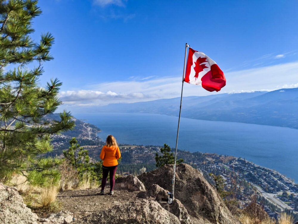 Gemma standing on the Pincushion Mountain summit looking at beautiful lake and lakeshore views on sunny day with Canadian flag flying adjacent