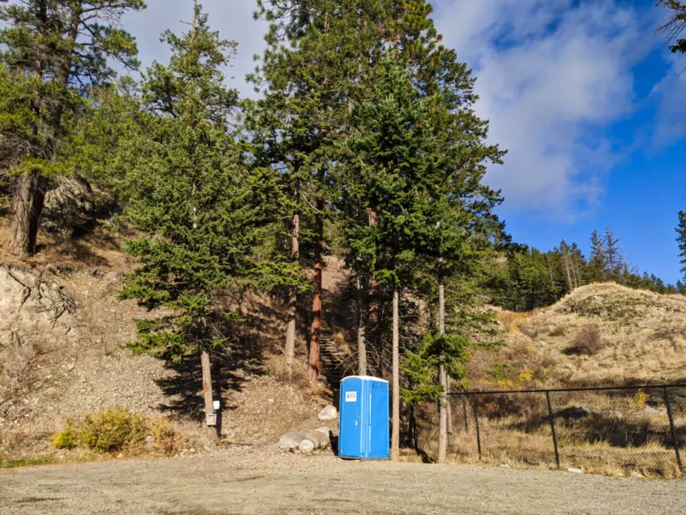 Looking across the Pincushion Mountain parking lot to blue porta potty standing next to trailhead with stairs visible behind