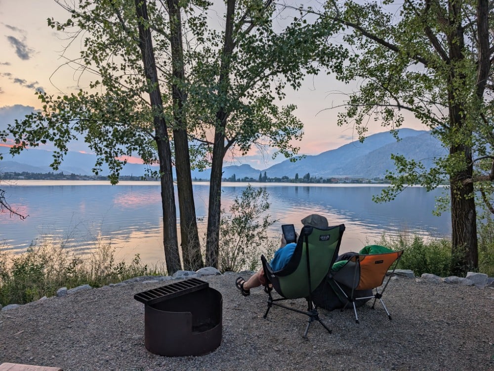 Back view of two camp chairs next to firepit (one occupied) in Haynes Point campground in Osoyoos, looking out to lake views