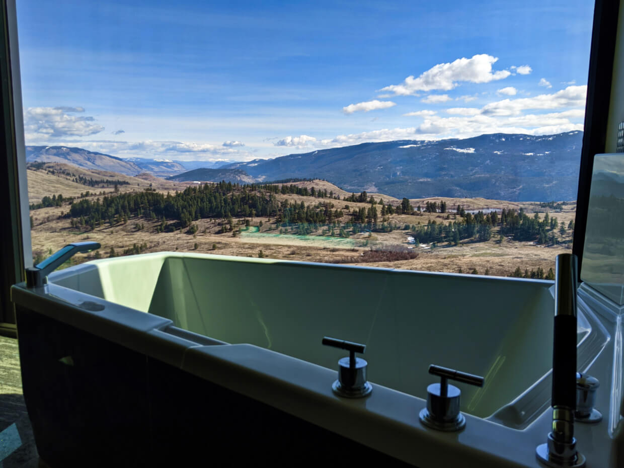 Two person bathtub in front of window with forest and hill  landscape shown through window behind