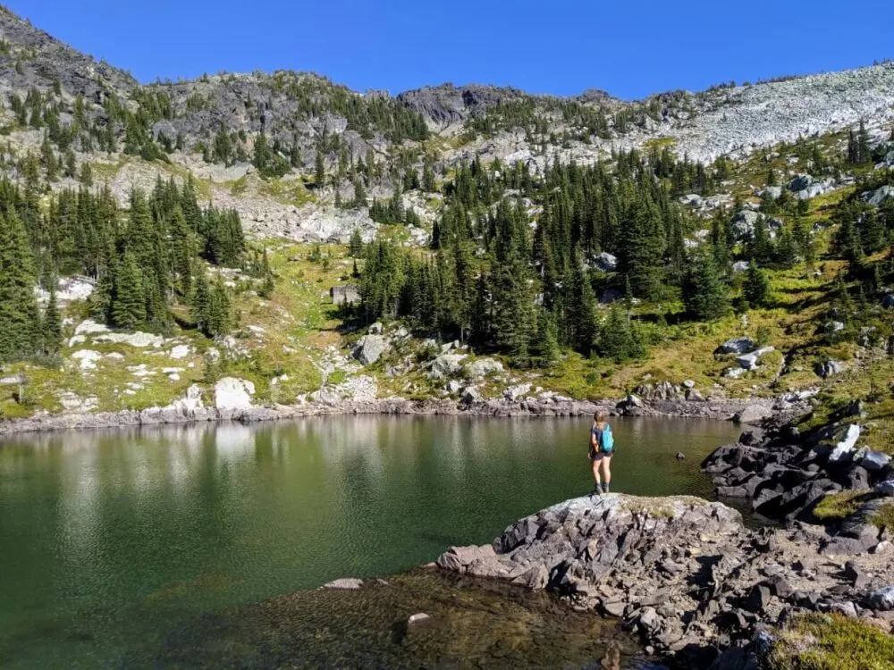 Gemma is standing on a rock jutting into lake, with forest and rock surroundings in background