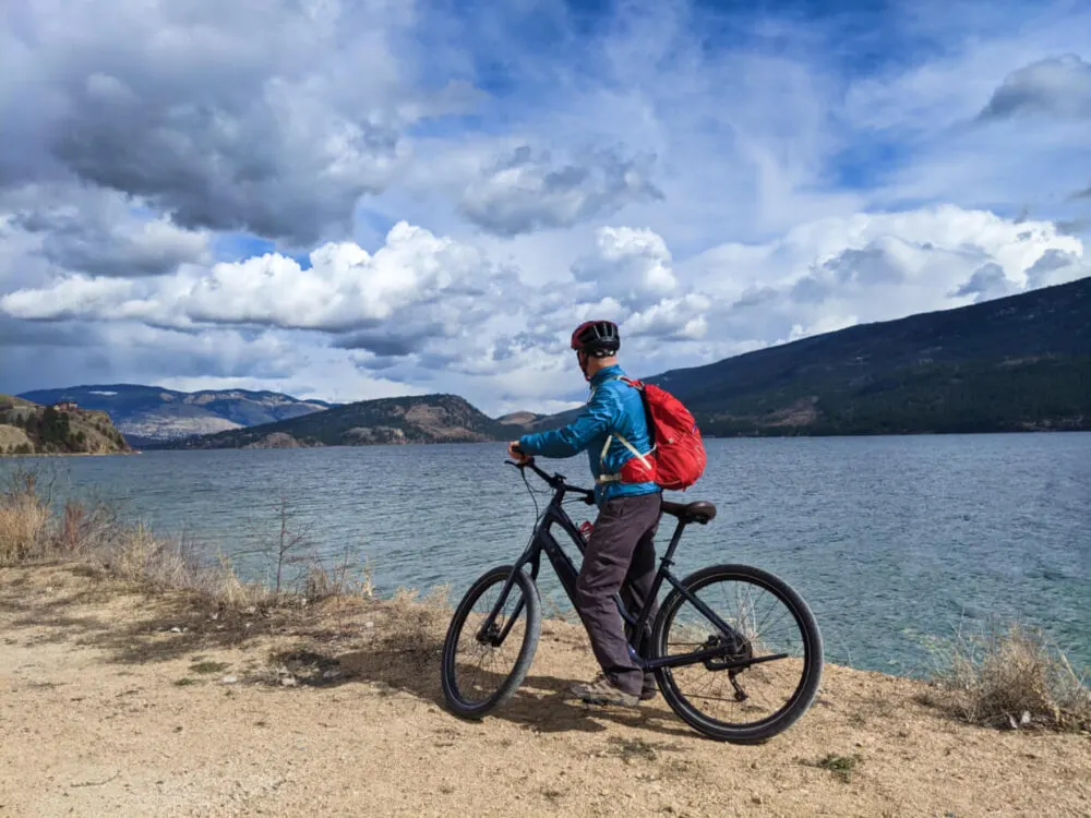 JR standing on electric assist cruiser bike next to Kalamalka Lake, looking towards hills in the distance