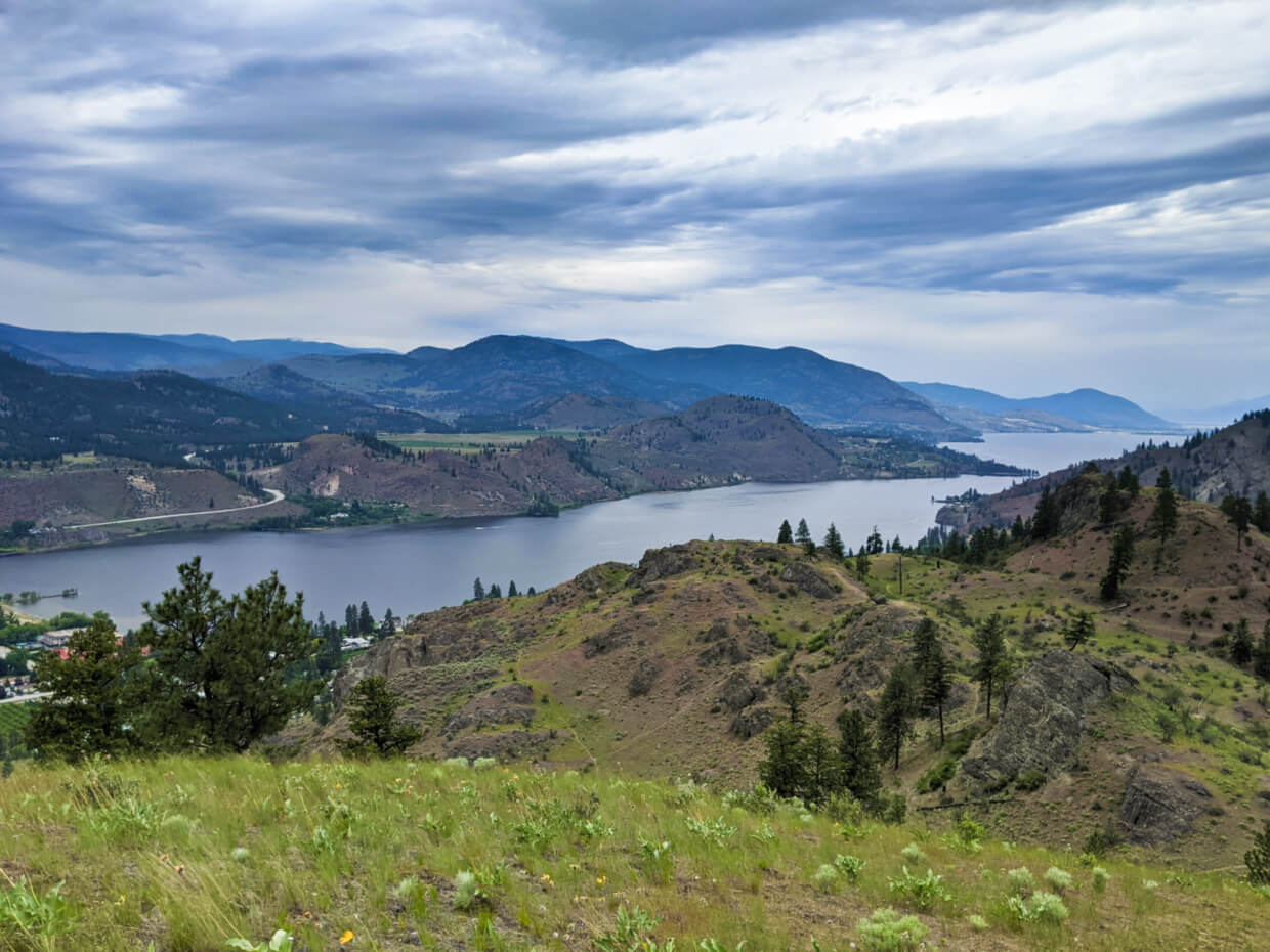 Looking out from elevated viewpoint across grassy hill with long lake in background, surrounded rugged hills and mountains