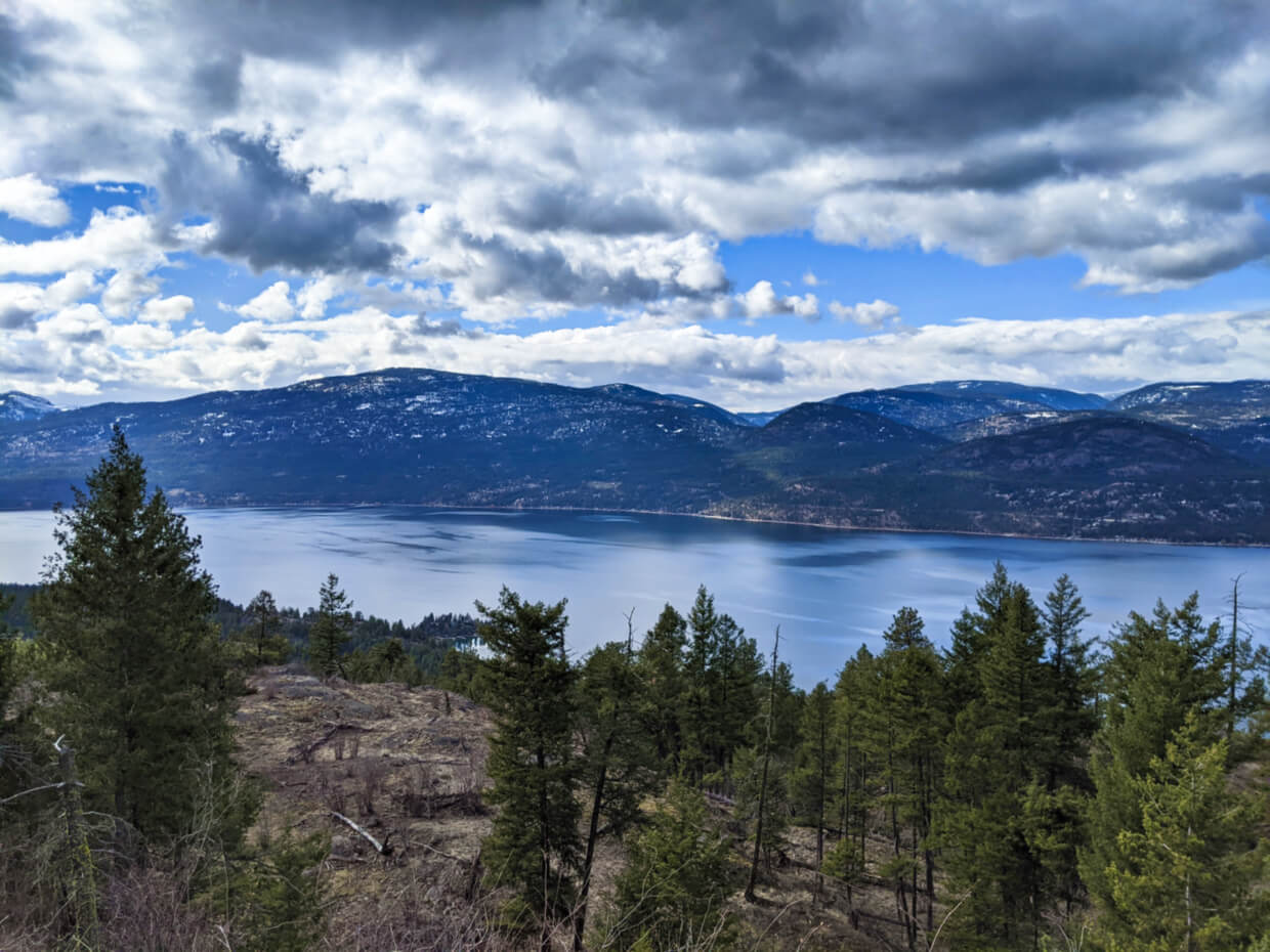 Looking down beyond trees towards Okanagan Lake, which is surrounded by mountains and hills, some with snow on the top
