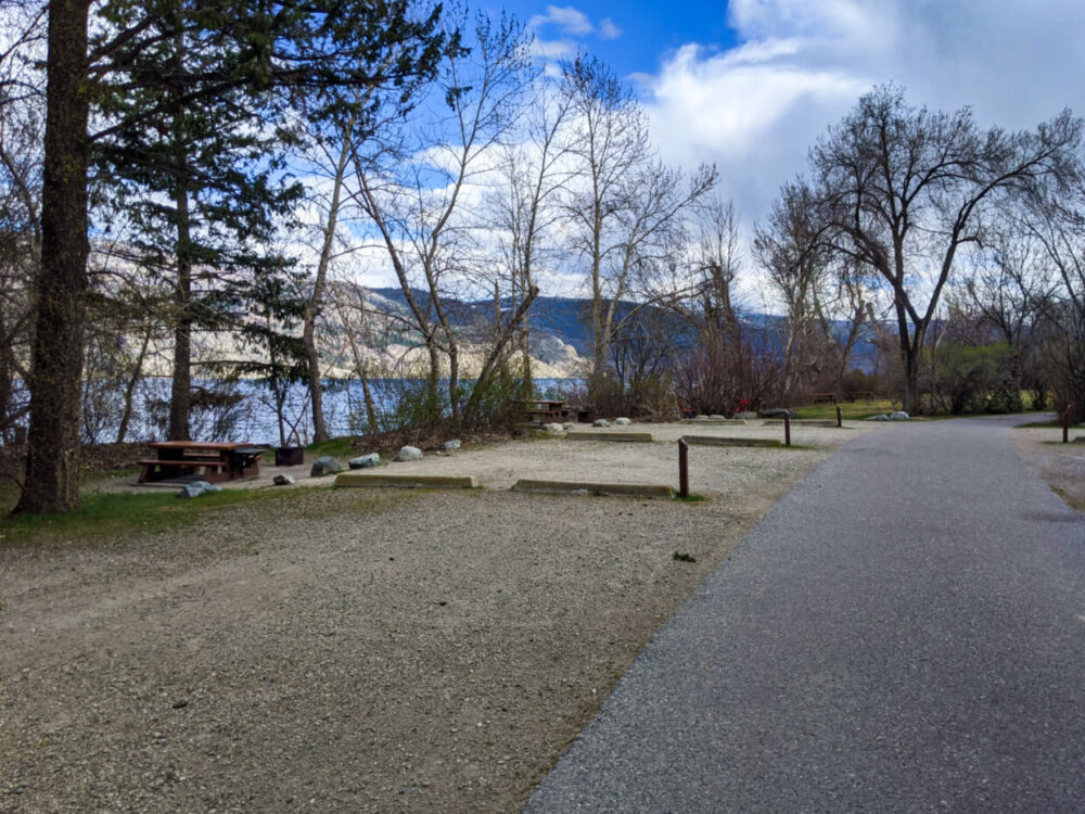 Side view of provincial park campsites, with large dirt parking area and picnic tables, located relatively close together. Okanagan Lake is visible in the background. 