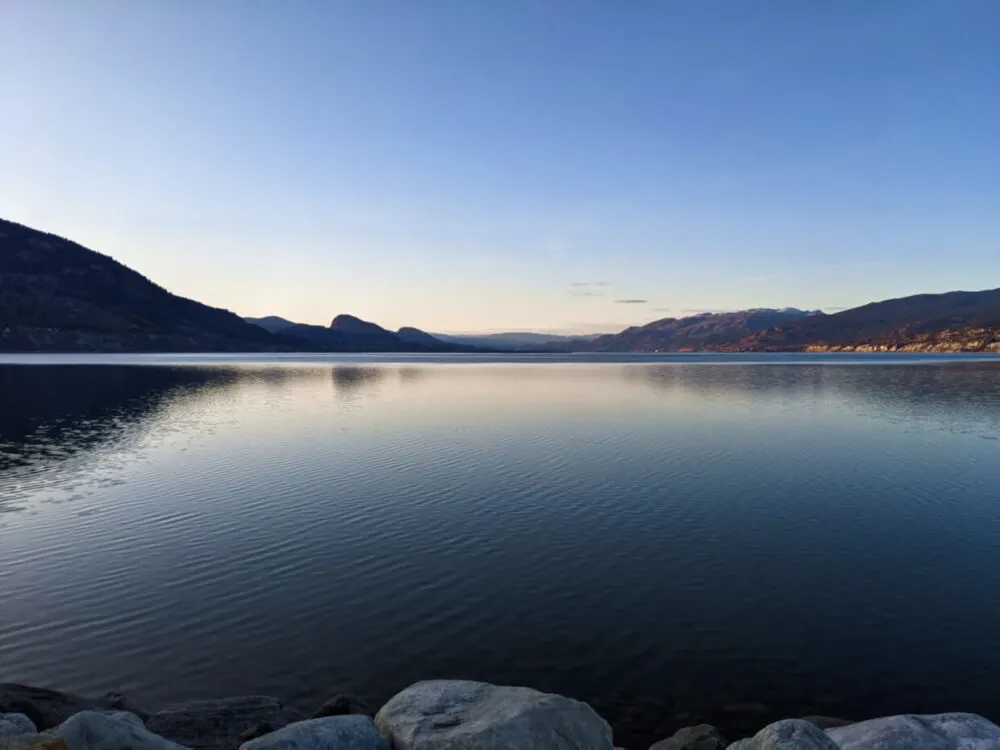 Lakeshore view of Okanagan Lake, with calm water and views of mountains behind