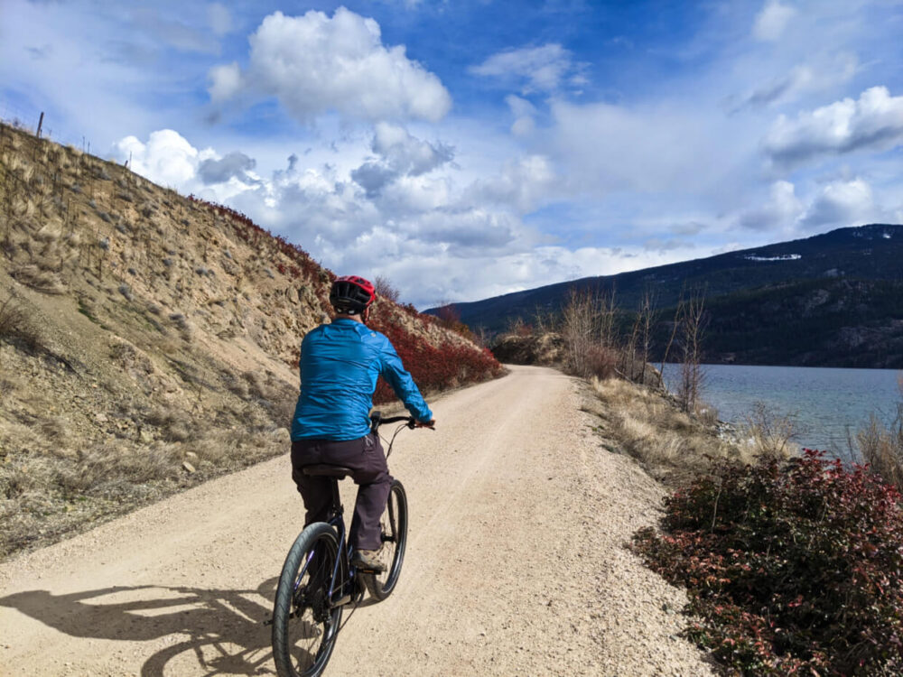 JR is riding e-cruiser bike on gravel trail next to Kalamalka Lake, with sloping hill on left