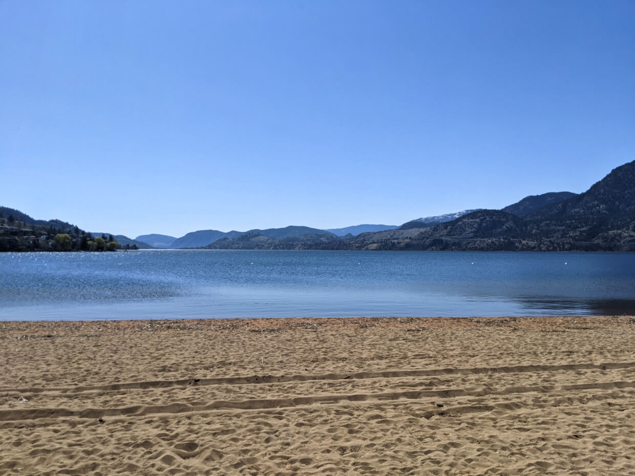 Looking out over the golden sands of Skaha Lake Beach to the calm surface of Skaha Lake, surrounded by mountains