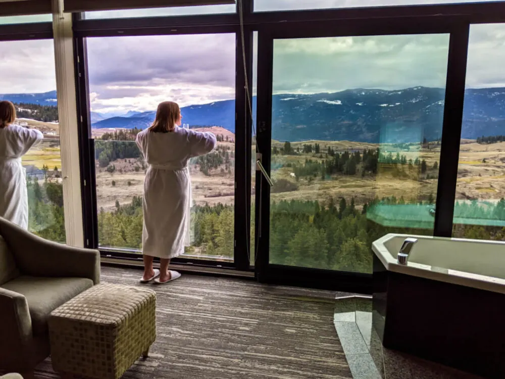 Gemma is standing by the floor to ceiling windows in Sparkling Hill hotel room, wearing a robe and looking out of the open window at views