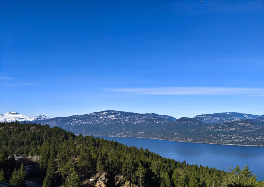 Looking out of the PeakFine restaurant windows to an elevated view of Okanagan Lake with hills behind, with one snow capped mountain in the distance. Forest is in the foreground