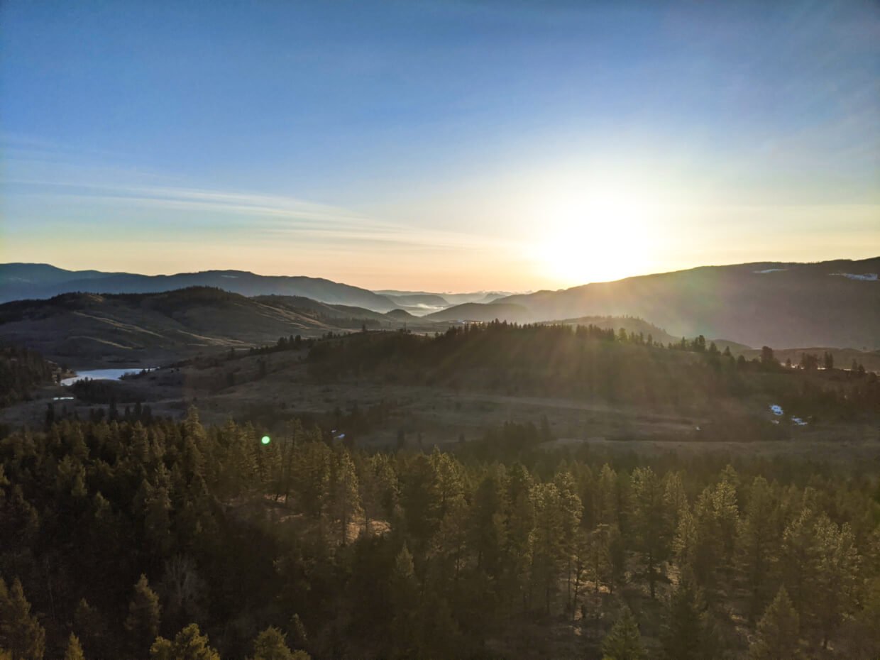 Elevated view of sun rising behind mountain with golden rays travelling towards camera, lighting up forest and grasslands landscape