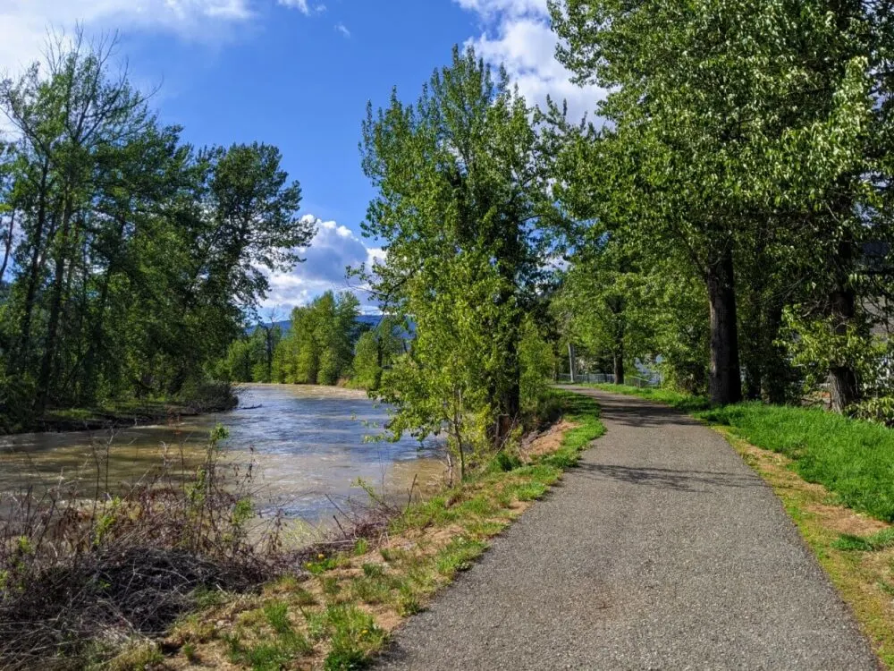 A paved trail (the Coldwater River Trail) runs alongside a rushing river, which is milky in colour. There are trees along the path