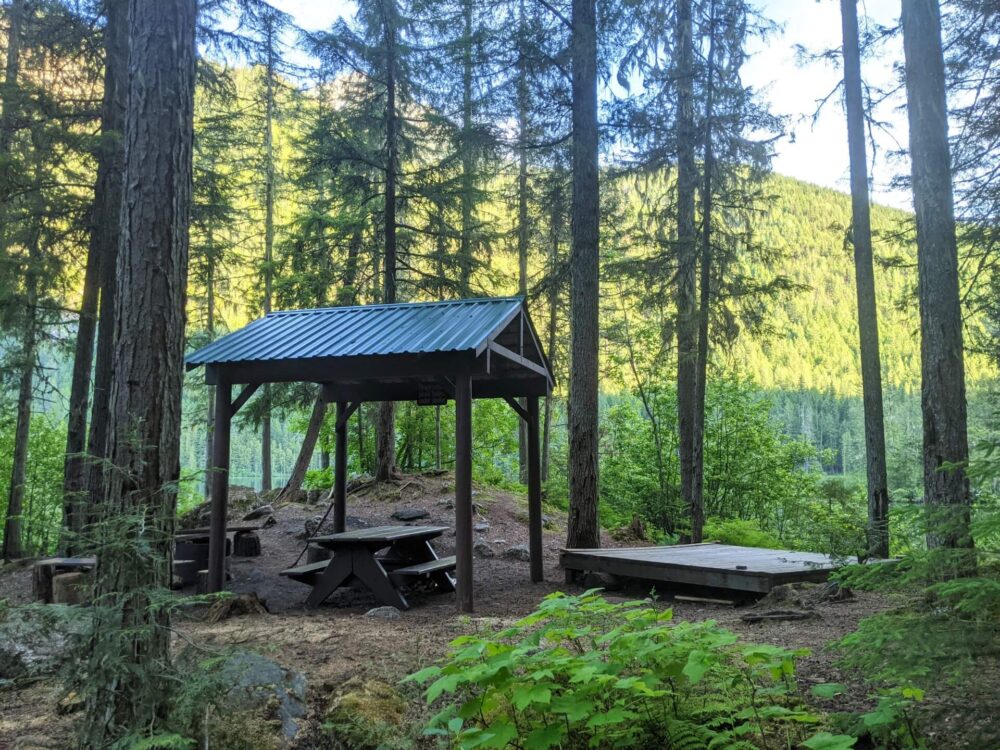 Distant view of wooden tent pad next to picnic table with shelter, with lake visible in the background