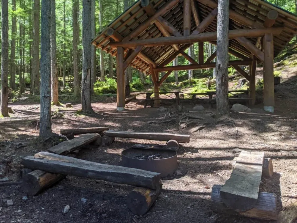 Looking across communal fit pit area with benches to large shelter with three picnic tables underneath in Spectrum Lake group campground