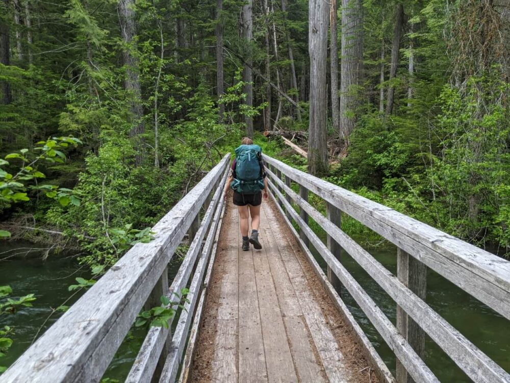 Back view of Gemma hiking away from camera on wooden bridge over river, carrying large turquoise backpack