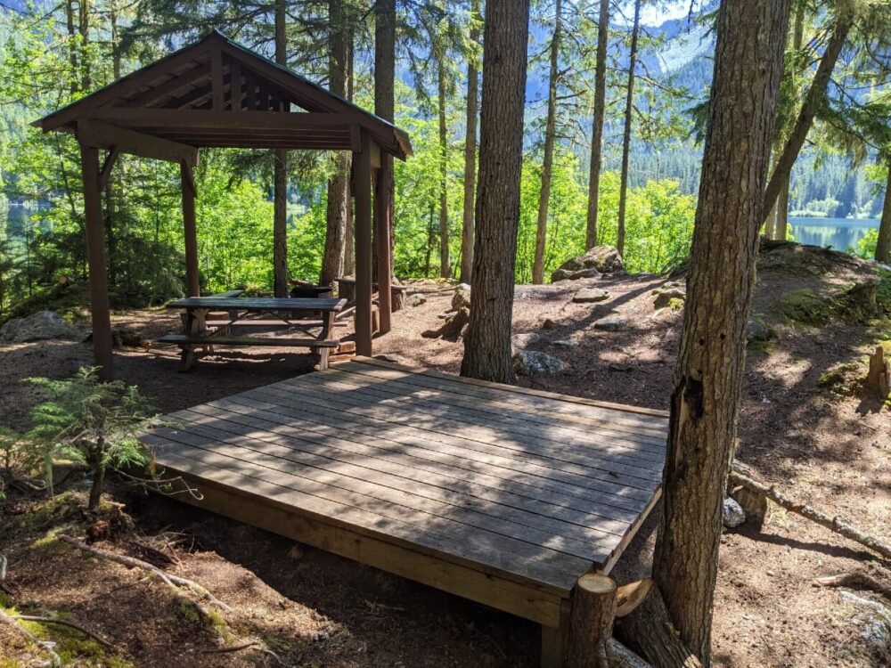 Raised wooden tent pad in front of picnic table with shelter, with lake visible in the background