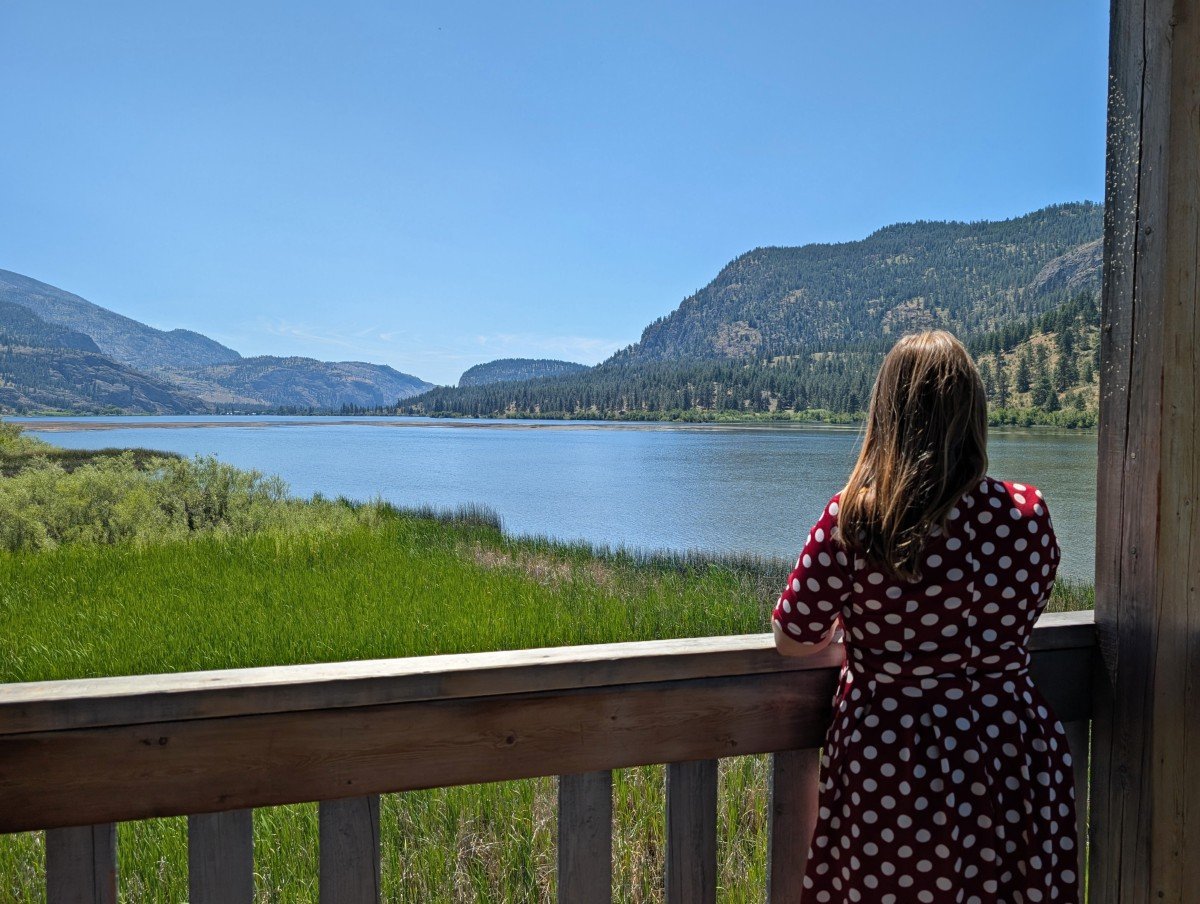 Back view of Gemma in red and white polka dot dress standing in Vaseux Lake bird hide, looking out to calm lake