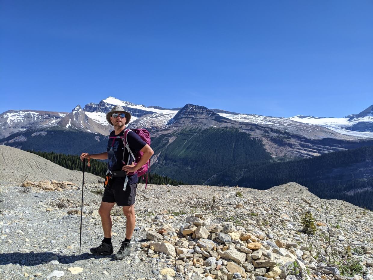 JR is standing and looking behind camera, with backdrop of rocky landscape with mountains and glaciers