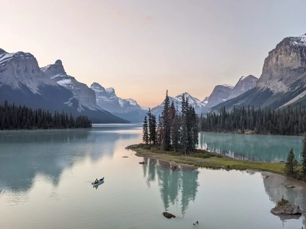 Looking across to Spirit Island (a peninsula with collection of trees), where a canoeist paddles across from the shore. Spirit Island surrounded by towering mountains