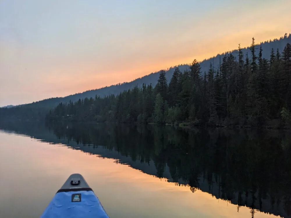 Sunset view of Jewel Lake from canoe, with orange colours behind mirror like reflections of lake and forest